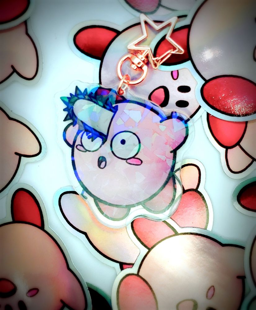 An acrylic charm of Chainsaw Kirby - a Kirby with a chainsaw sticking out of their head. They look surprised. The charm has a "cracked" holo finish.