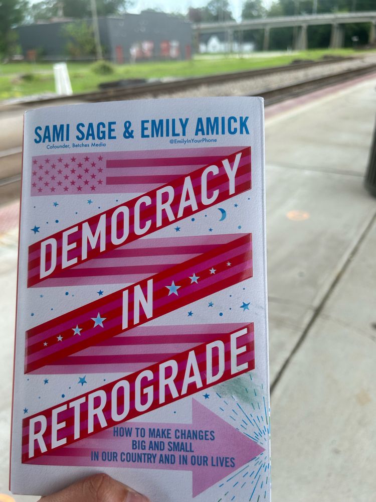 Book slightly off center in front of railroad tracks. The title of the book is in bold letters: Democracy in Retrograde by Sami Sage & Emily Amick.