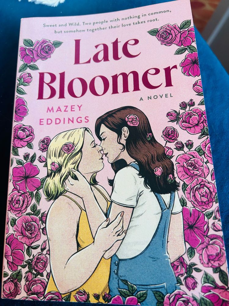 Photo of the cover of Late Bloomer by Mazey Eddings featuring a short blonde woman a moment away from kissing a tall brunette woman in overalls. They are surrounded by gorgeous pink blooms. 