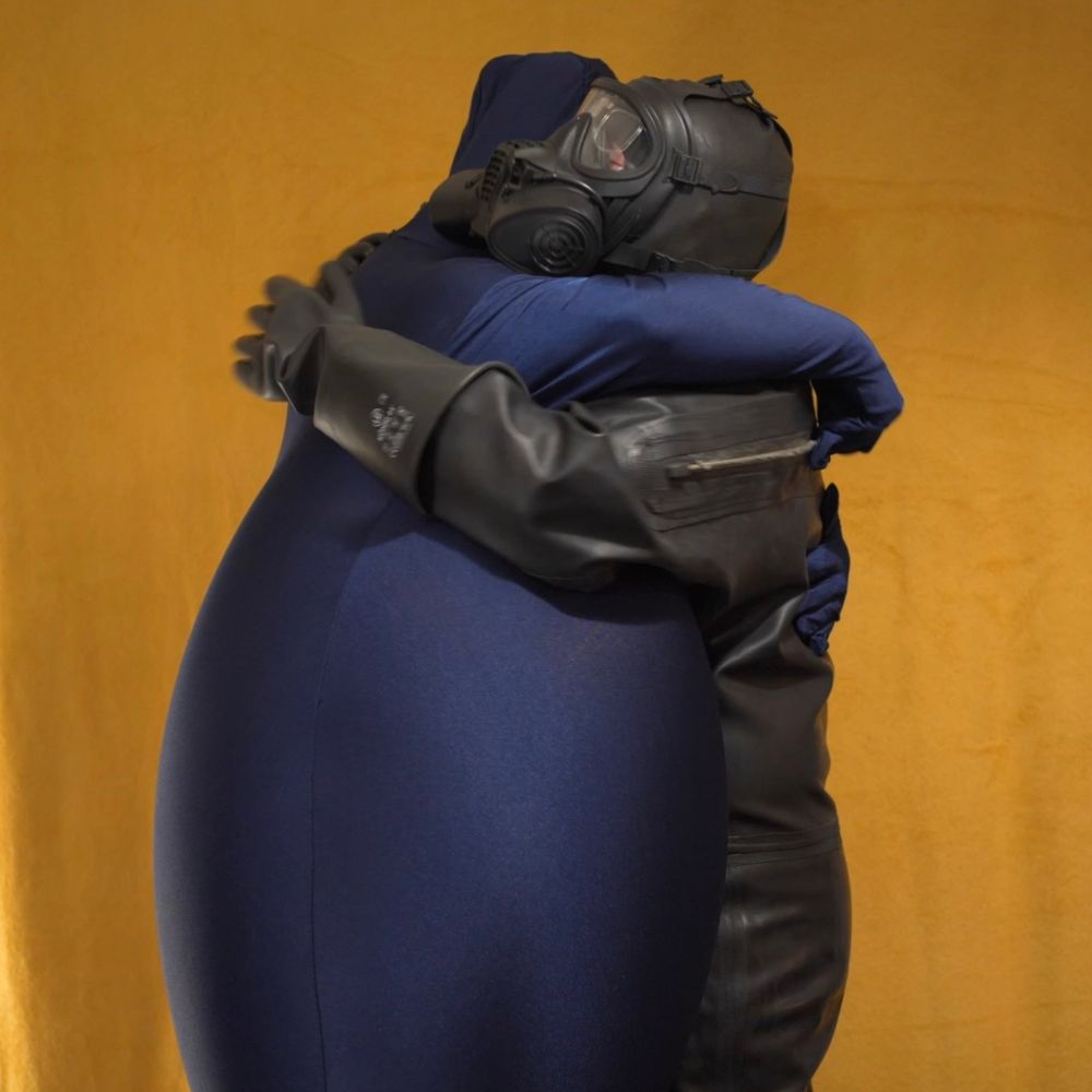 Two masked drones stand in front of a yellow background. The drone on the left is in an overly inflated blue zentai suit, the one on the right is in a black rubber drysuit, they both share a loving puffy embrace in each others arms. 
