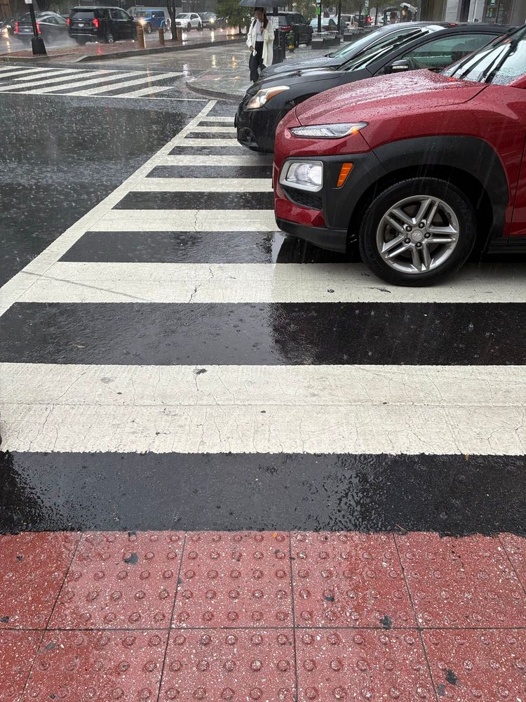 A rainy crosswalk with three cars abreast, all protruding deep into the crosswalk so pedestrians can hardly cross safely