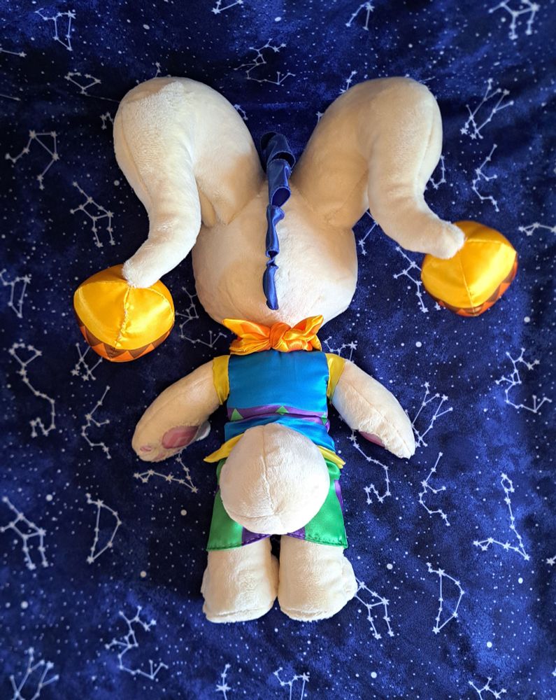 A photo of a handmade plush toy of Misfit, a cream-toned bunny-like plushcritter with green button eyes, a zipper-style mouth made of satin, and long ears with bells on the ends. He is made mostly of minky and satin. This photo shows his back side, including a large round tail.