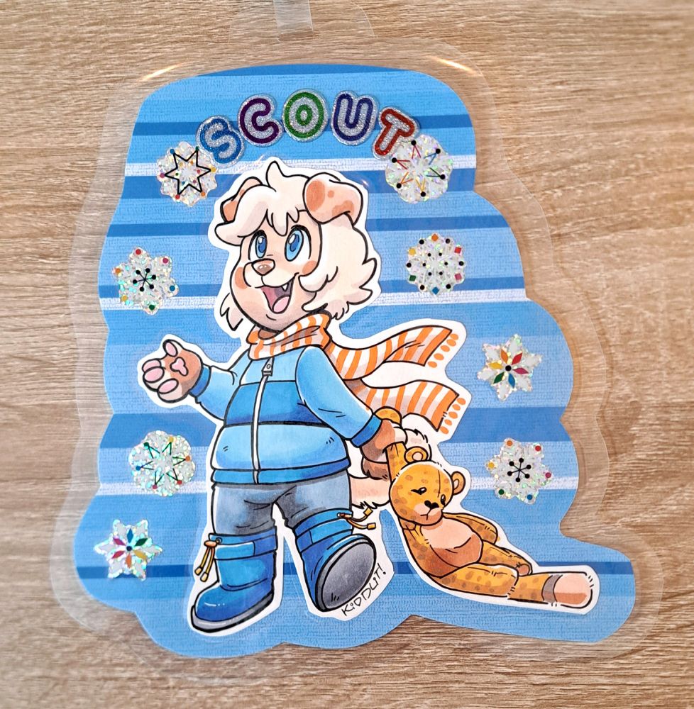 A photo of a marker badge featuring Scout, a kidfur puppy, wearing winter gear, with his cheetah plushie. There are snowflake stickers surrounding him on a patterned blue textural paper and the name Scout spelled out in bubble stickers.