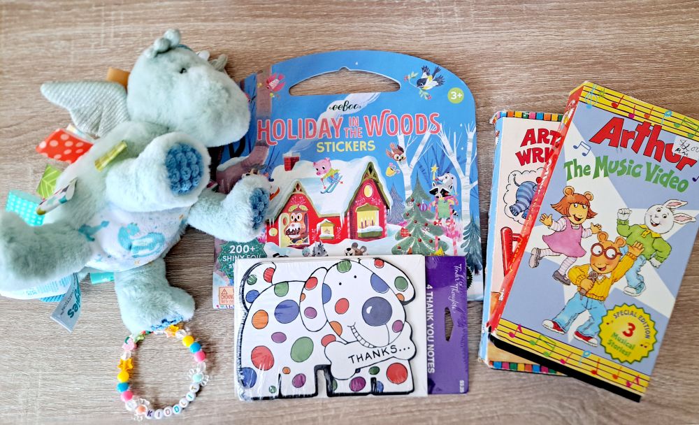 A photo of a Taggies dragon plush, kandi bracelet, vintage spotted puppy thank you cards, a holiday sticker book, and retro Arthur vhs tapes.