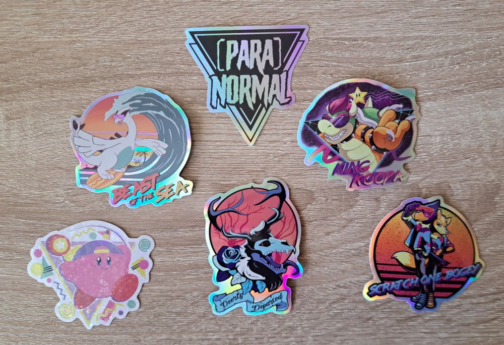 A variety of artist stickers - an upside down triangle reading ParaNormal, a surfing Lugia sticker, a retro Bowser sticker, a 90s style yo-yo Kirby sticker, a Deerly Departed skull monster cryptid sticker, and a retro style Star Fox and Falco sticker.