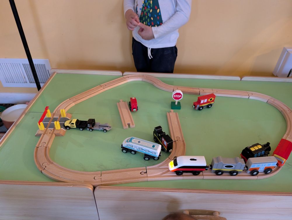 A little boy stands behind a train table. An electric locomotive pulls two cars around a track. Two hot wheels trucks wait at a train crossing. An offshoot of the track leads to a stop sign 