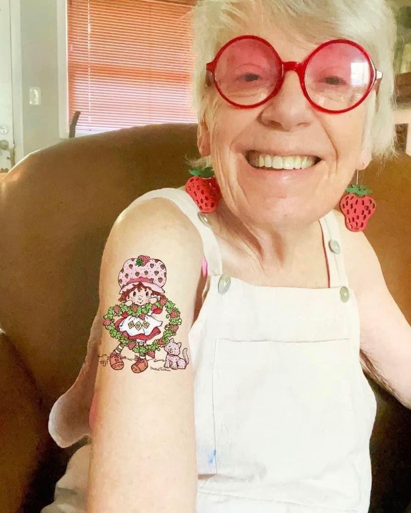 A photo of Muriel Fahrinon, creator of the Strawberry Shortcake and Care Bears. An older woman with white hair, She has red rimmed glasses, strawberry earrings and a strawberry shortcake tattoo. She is smiling. 