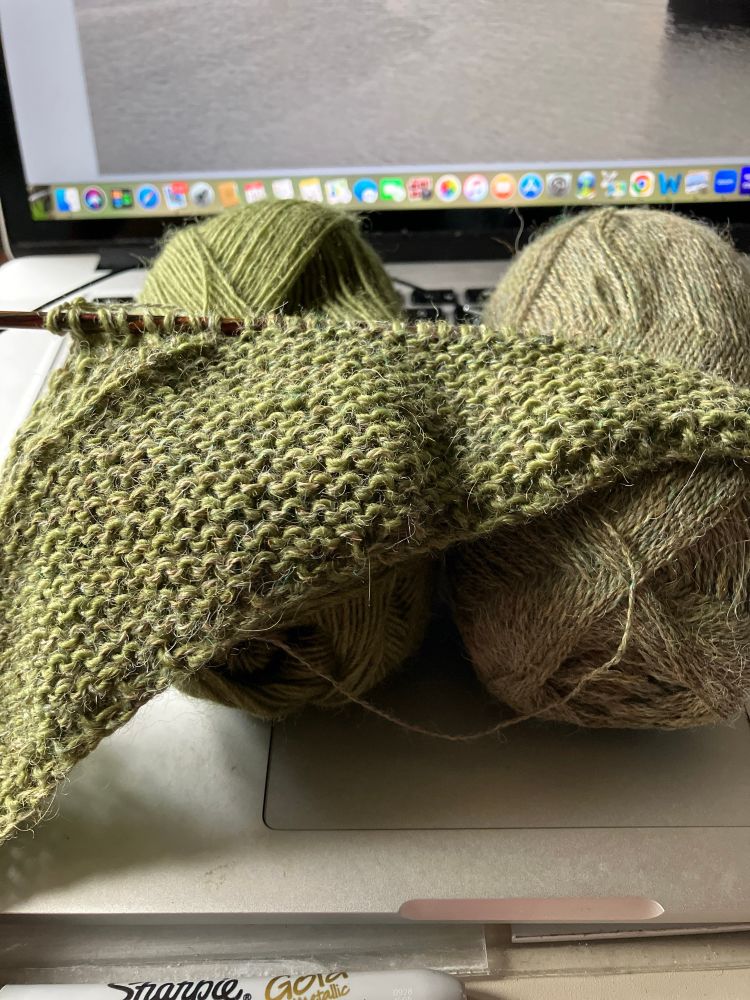 A triangular piece of knitting sits on top of 2 balls of green yarn. The knitter has put it down for a break while she makes a cup of tea.