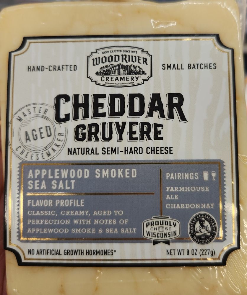 A squarish block of white cheese labeled "CHEDDAR GRUYERE natural semi-hard cheese". Other text on the label includes: Proudly Wisconsin Cheese, master cheesemaker aged, hand-crafted, small batches, Wood River Creamery, no artificial growth hormones*, net wt 8oz (227g), and perhaps most offensively to both the English and the Swiss, "Applewood smoked sea salt" appears to be a flavor, and it is suggested to be paired with farmhouse ale and Chardonnay. 
