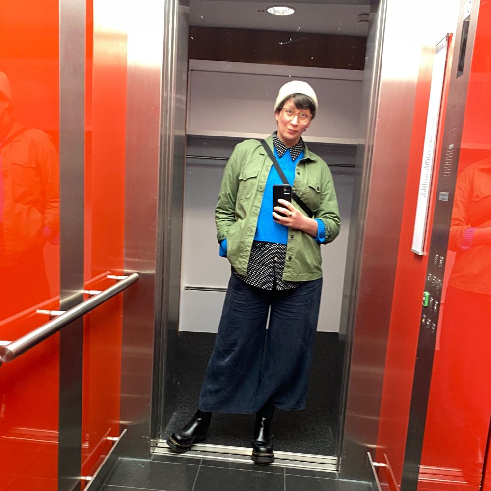 Person doing a selfie inside an elevator with dark pants, a polka dot blouse layered under a blue sweater and a green jacket. 