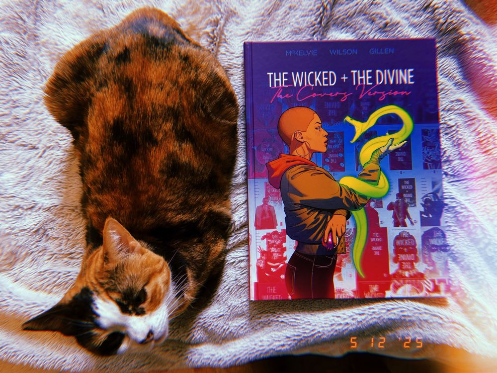 photo of a calico cat sitting with a wicked and the divine covers version book on a furry blanket. 