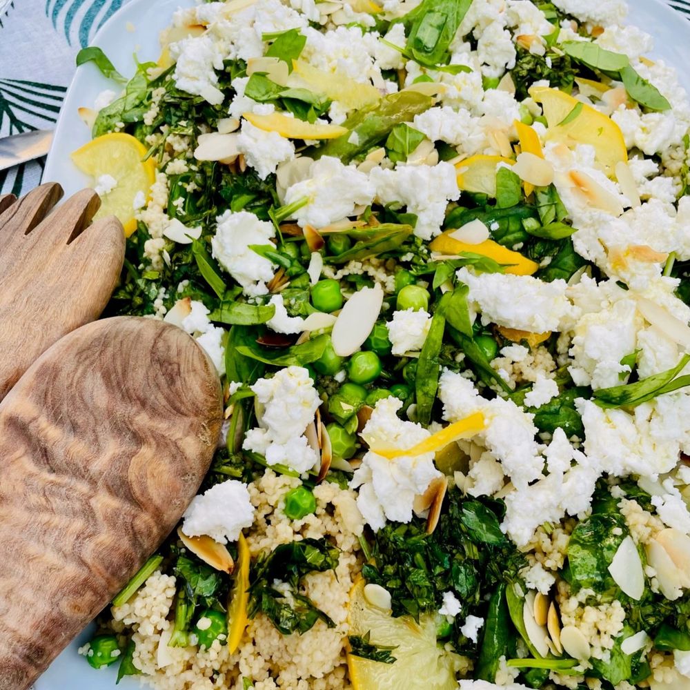 Salat mit Couscous, Erbsen, Salzztrionen, Feta und kleingehacktem Spinat auf weißem Teller, daneben Salatbesteck aus Olivenholz.