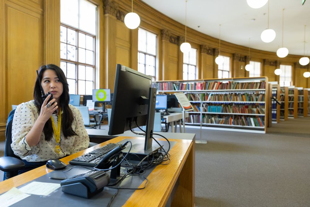 A woman speaksn on a walkie talkie in a library whilst looking at a computer screen.