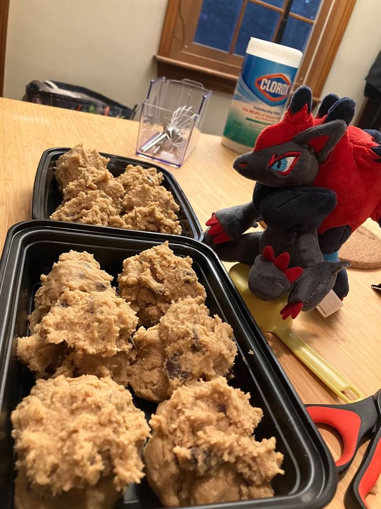 A kitchen table with two containers of cookie dough measured out. Sat nearby on a measuring cup is a plushie zoroark that I call my son.