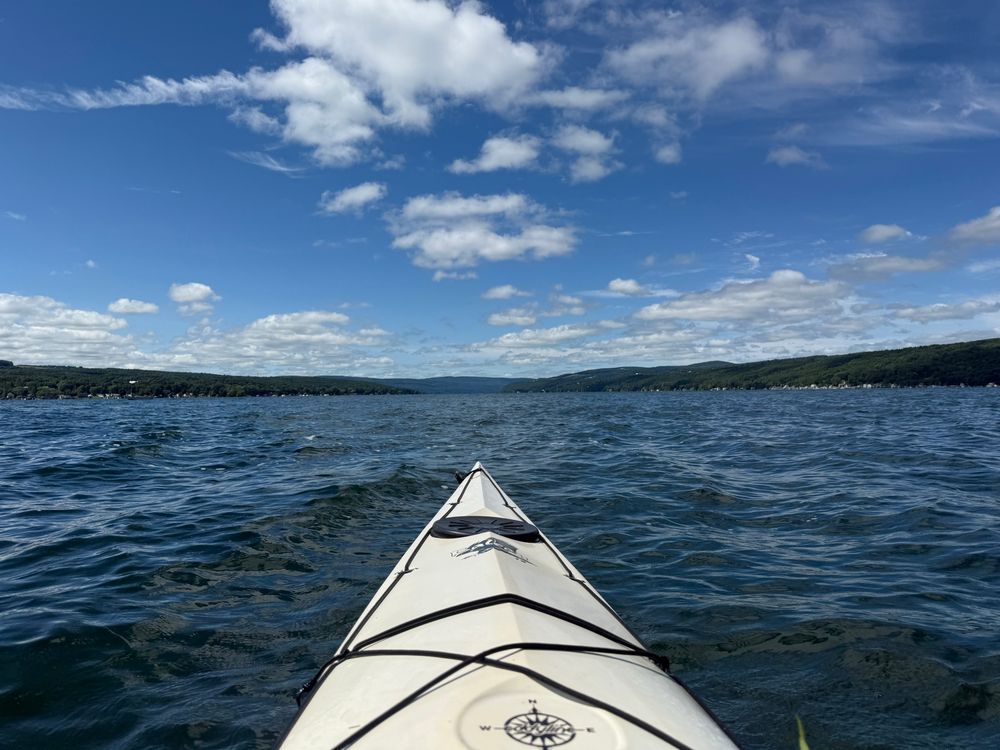 The nose of a white 18’ sea kayak in deep blue waters with a lighter blue sky sporting fluffy white clouds 