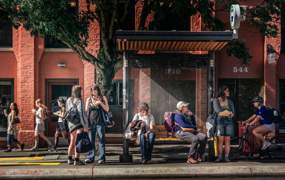 Tableau of people at or around a bus stop in Victoria, BC, on a sunny afternoon.  There is a trio of young girls eating snacks on the left, a pair of woman chatting to their right, three women apparently waiting at the bus stop itself, and a woman passing on a bike on the far right.  There are a brick building and a fairly large urban tree in the background.