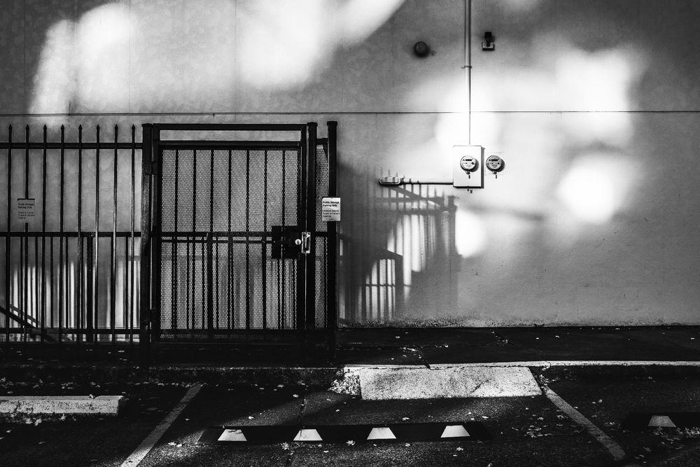 B&W parking lot scene with sunlight and shadows cast upon a white featureless building.  There are the front of parking spots along bottom, and a fence/gate on the left middle.