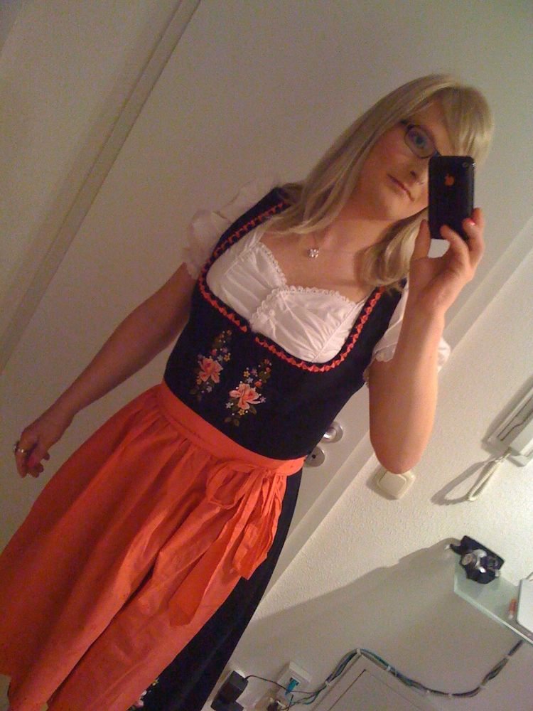 Eine Person mit schulterlangem blondem Haar und Brille macht ein Selfie in einem Spiegel. Sie trägt ein traditionelles Dirndl mit einer weißen Bluse, einem schwarzen Mieder mit bunter Stickerei und einer roten Schürze. Die Person hält ein schwarzes Smartphone in der rechten Hand, während ihr linker Arm leicht ausgestreckt ist. Der Hintergrund zeigt eine weiße Wand mit einer Gegensprechanlage und einem kleinen Spiegelregal mit ein paar Gegenständen darauf.