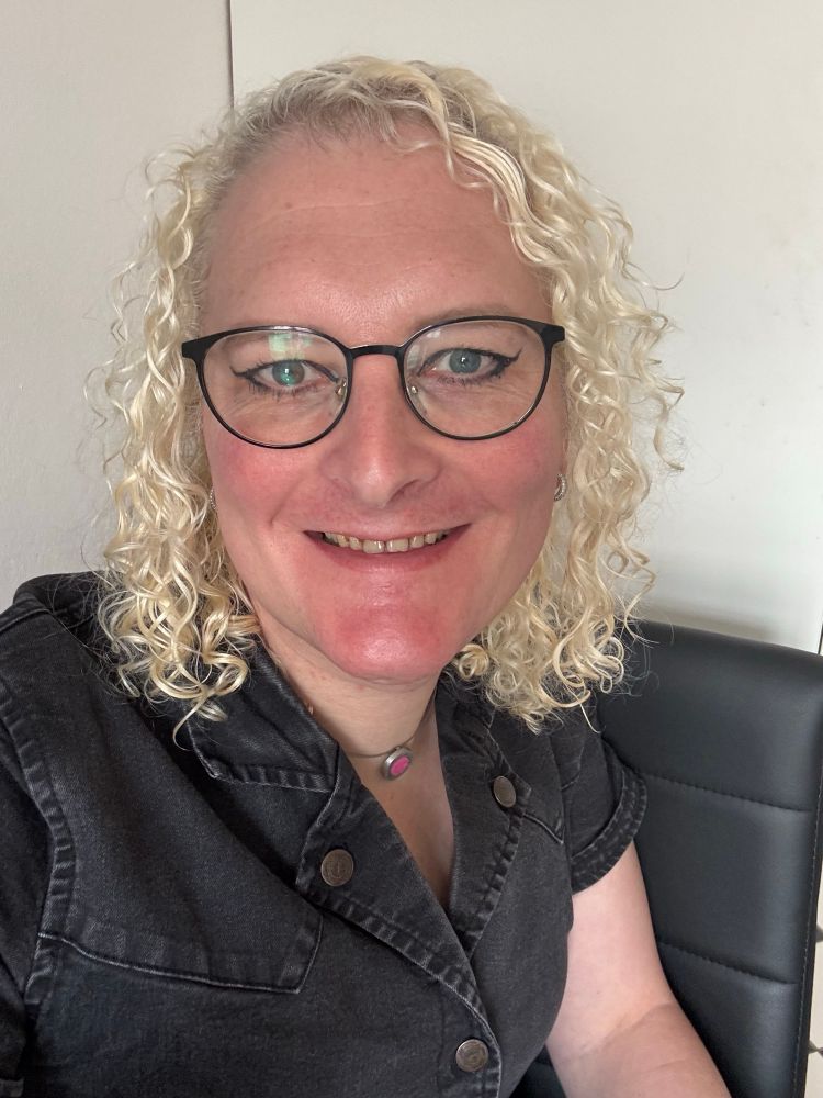 Eine lächelnde Frau mit blonden, lockigen Haaren und schwarzer Brille sitzt auf einem schwarzen Stuhl vor einer weißen Wand. Sie trägt ein dunkles Jeanskleid mit Druckknöpfen und einen Anhänger mit rosa Filz an einer Silberkette. Ihr Teint ist hell, sie trägt dezent Make-up mit rosigem Lippenstift. Das Licht ist natürlich und hell, das Bild vermittelt eine freundliche und offene Stimmung.