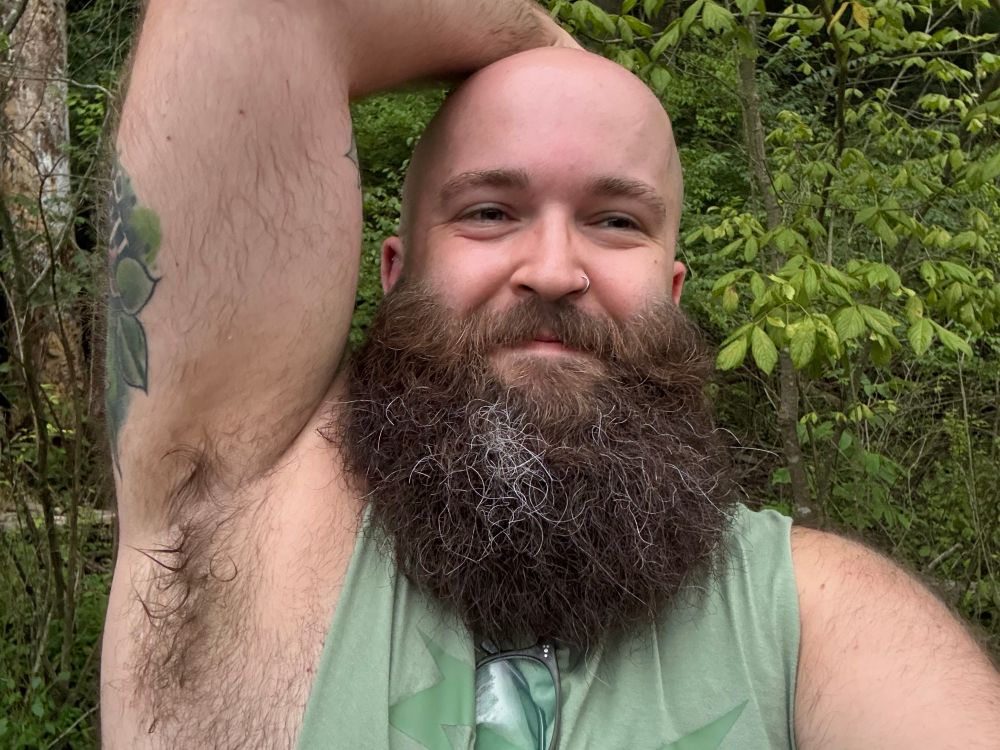 A selfie of the author of this post (a bald nonbinary person with a bushy brown beard in a cutoff shirt) showing off their sweaty armpit in the woods.