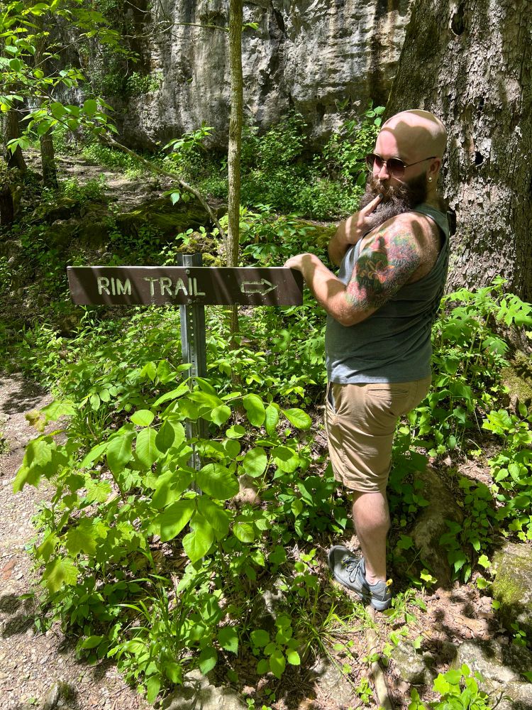 The author of this post standing in nature beside a sign that says "rim trail" with an arrow pointing to the right at the author. The author is facing away from the camera so their butt is visible and glancing over their shoulder provocatively.