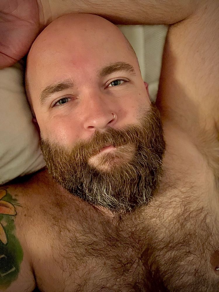 The author of this post, a bald white person with a dark brown beard, looking at the camera while laying naked in bed. They are only visible from the mid-chest up.