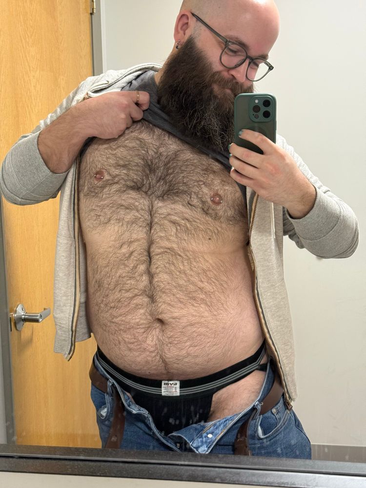 A selfie of me in a mirror. I've pulled my shirt up so you can see my hairy chest and belly. I've also pulled down my shorts enough that you can see I'm wearing a black jockstrap.