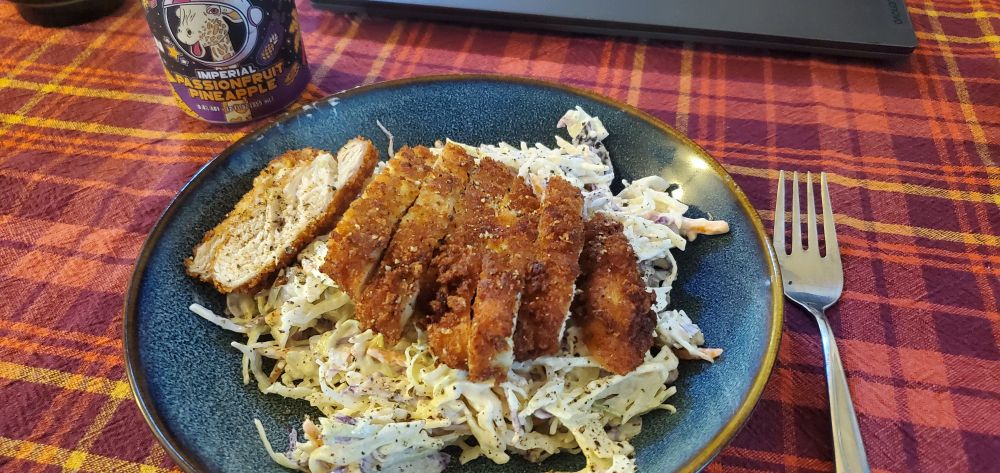A bed of multicolor slaw with a creamy dressing topped with a sliced piece of chicken katsu on a blue bowl-plate. It's on an autumnal tablecloth with a can of passionfruit pineapple cider