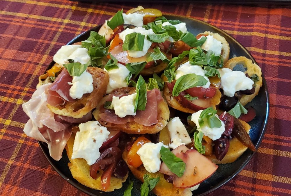 A plate piled high with stone fruit bruchetta topped with chunks of burrata and torn basil. Several also have pieces of prosciutto 