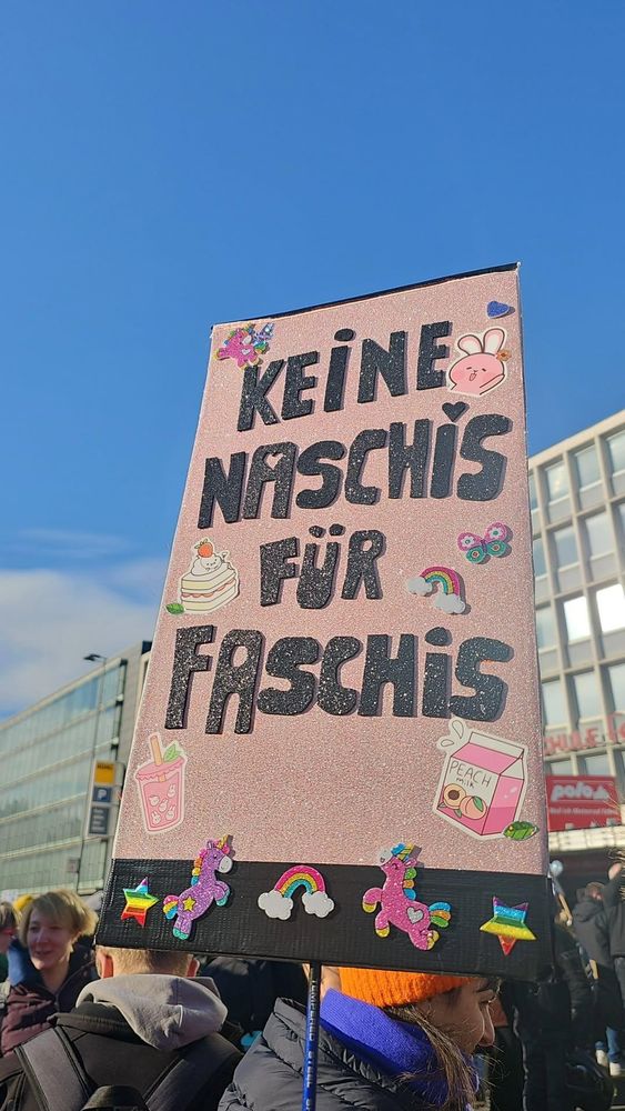 Ein rosa glitzerndes Schild vor einem blauen Himmel und grauen Gebäuden mit Menschen im Hintergrund. Auf dem Schild steht in schwarzer Glitzerschrift "Keine Naschis für Faschis", verziert mit Aufklebern 