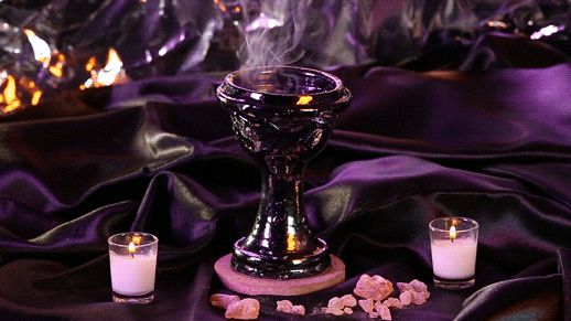 Incense smoke coming out of a fancy goblet, surrounded by candles, crystals, and purple folded fabric.