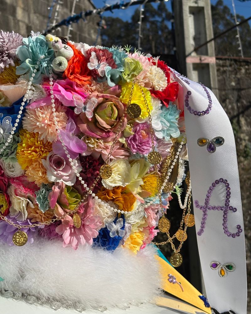 Un chapeu de Madama do Entroido galego, decorado cunha gran variedade de flores de cores, perlas, cadeas douradas con moedas e bolboretas brillantes. A un lado colga unha banda branca coa letra “A” feita con pedrería e pequenos adornos. A base do chapeu está rematada cun ribete de pelo branco. Ao fondo vense luces festivas colgadas e elementos de pedra nun entorno exterior asollado.