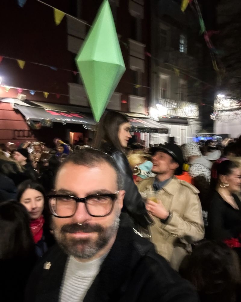 Fotografía dunha rúa durante unha festa nocturna (Entroido) chea de xente disfrazada. No primeiro plano, un home (eu) con lentes e barba sorrí lixeiramente á cámara, leva na cabeza un obxecto verde en forma de diamante, similar ao “plumbob” dos videoxogos The Sims. A escena está iluminada por luces e decoracións festivas con bandeirolas de cores.