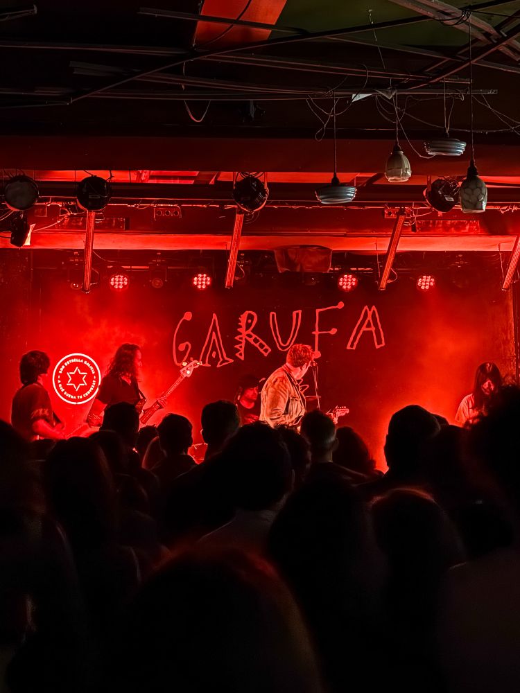 A live concert at Garufa, a music venue. The stage is illuminated with intense red lighting, and The Pains of Being Pure at Heart perform in front of a crowd. The silhouettes of the audience contrast with the bright stage, where the musicians, including a guitarist and a bassist, are immersed in the music. 