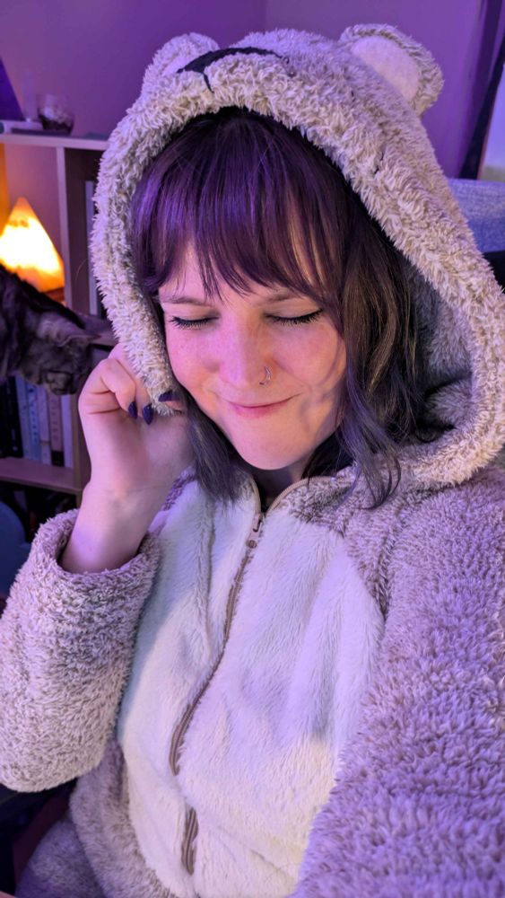 A selfie of the streamer wearing a cosy teddy bear onesie with the hood up. She is smiling at the camera with her eyes closed. There is a cat in the background and a salt lamp.