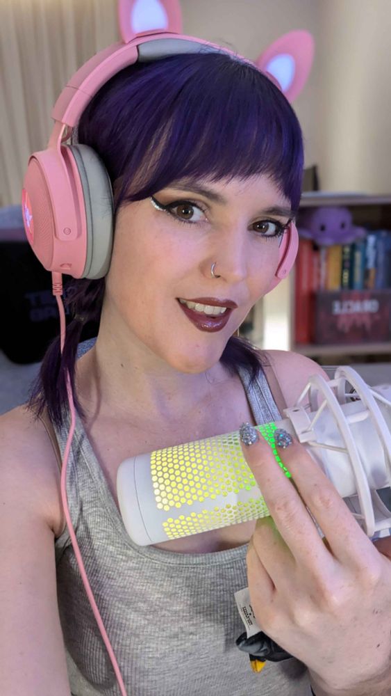 A photo of the streamer smiling at the camera holding an RGB microphone in front of her. She has purple hair in two small low plaits. She is wearing pink Razer headphones with light-up cat ears. She has winged eyeliner and sparkly finger nails. She is wearing a grey tank top.