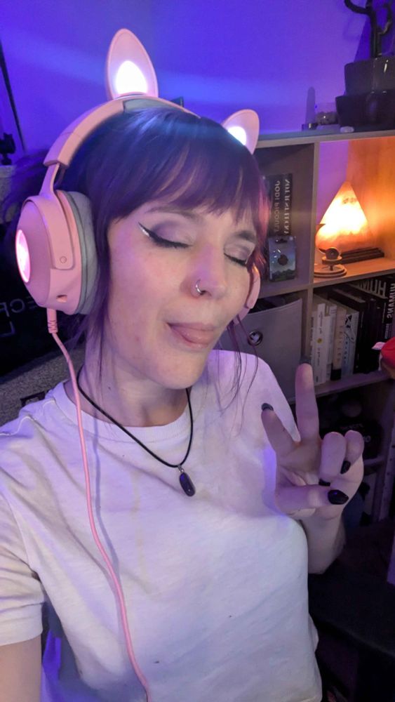 Peets wearing Razer headphones with LED cat ears while holding two fingers up in the peace symbol. She has her eyes closed and tongue sticking out . She is wearing a white t-shirt and has winged eyeliner.