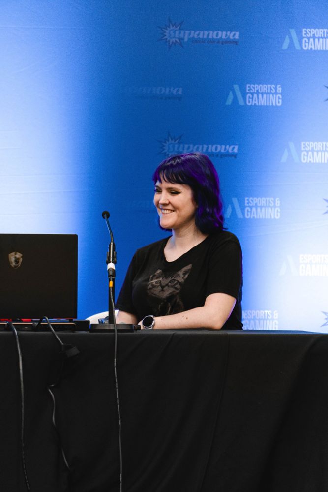 Peets is sitting at a table with an open laptop and a microphone in front of her. She is smiling and playing a video game. She has purple hair and is wearing a black t-shirt with a cat on it. The table is covered in a black table cloth. The backdrop is blue and features logos from Supanova logos from the event, and Aus E Spots logos from the panel sponsor.