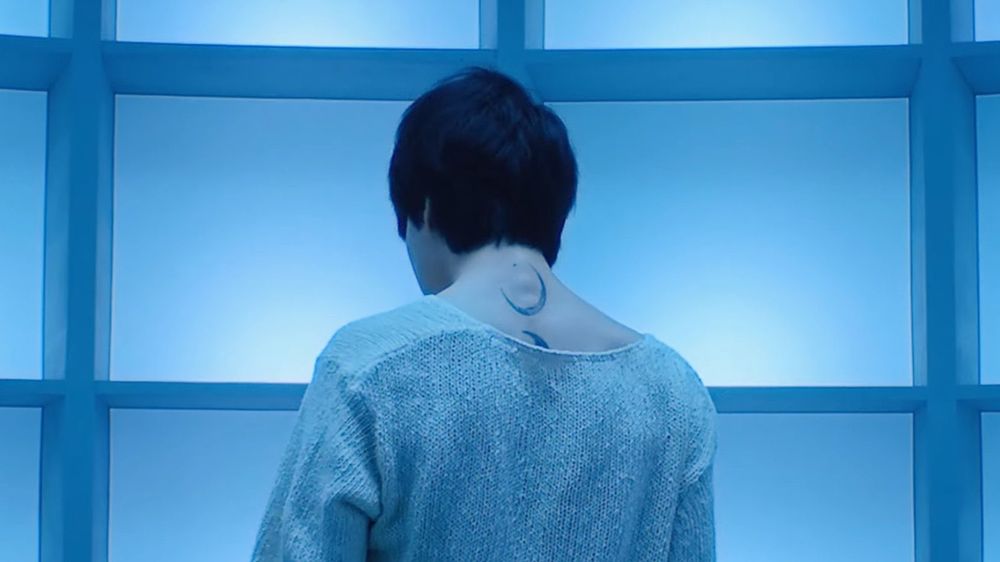 A picture of Jimin from the Set Me Free Pt 2 music video. He is facing away from the camera wearing a loose white sweater. You can see his moon tattoos. 