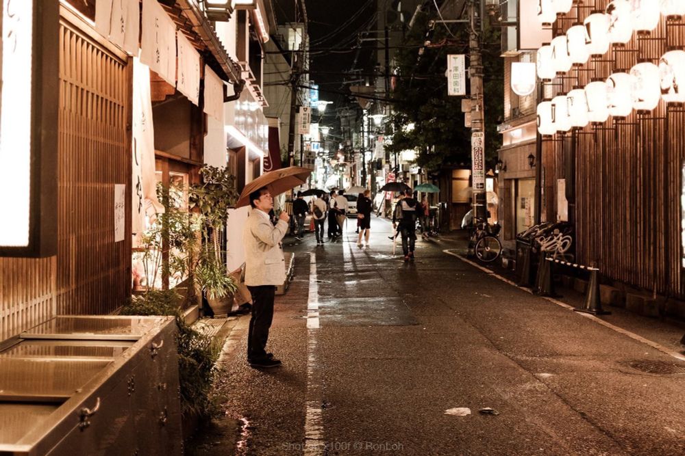 A man holding an umbrella staring into a street lighted up by rows of orange lanterns. 