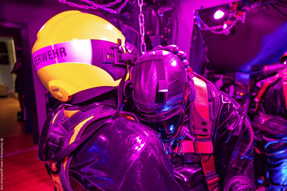 Two rubber drones hugging each other, one in a German firefighter helmet, and the other wearing a fall protection harness, a MSA gas mask, and a Loitokari rubber drysuit.