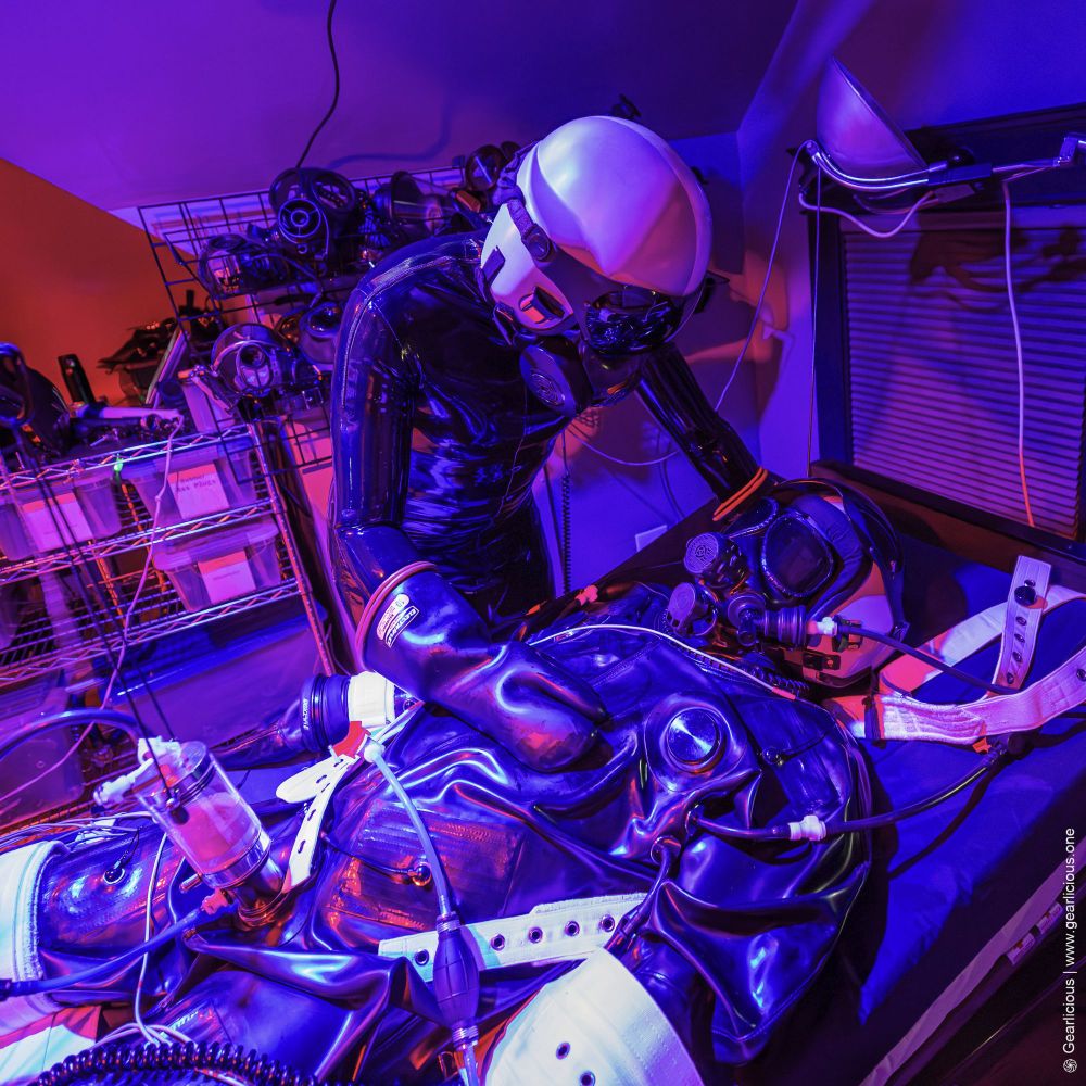 Rubber drone standing over another that's bound on a bed and wearing a vacuum rubber suit in Segufix. They are wearing gas masks and matching helmets.
