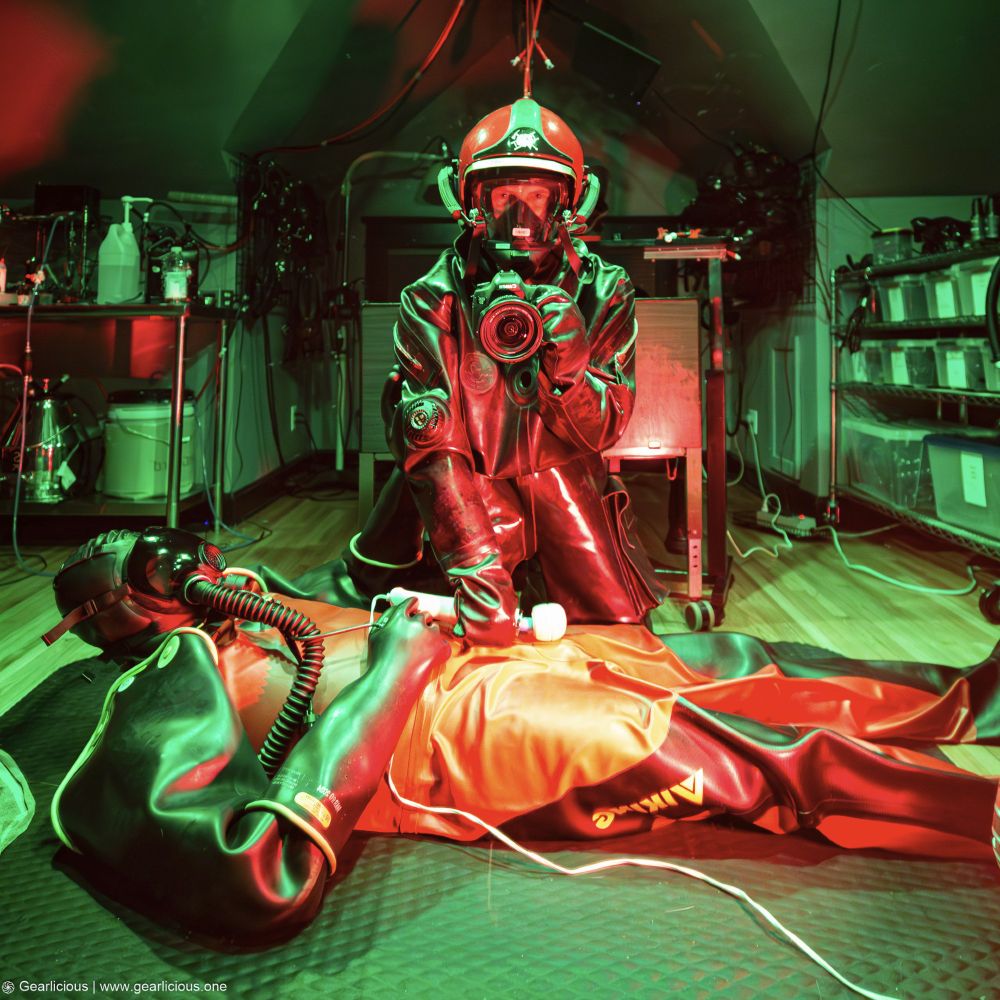 Two rubber drones in heavy rubber drysuits, one taking the photo in a european firefighter helmet and gas mask holding a vibrator up to another drone in a drysuit and a MSA gas mask that is laying on the floor.