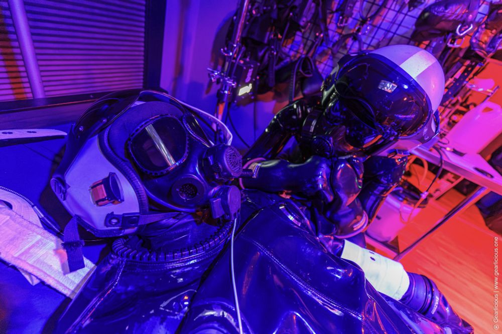 Two rubber drones wearing identical helmets, in gas masks. One is bound to a bed, and another is staring down at them in a menacing pose.