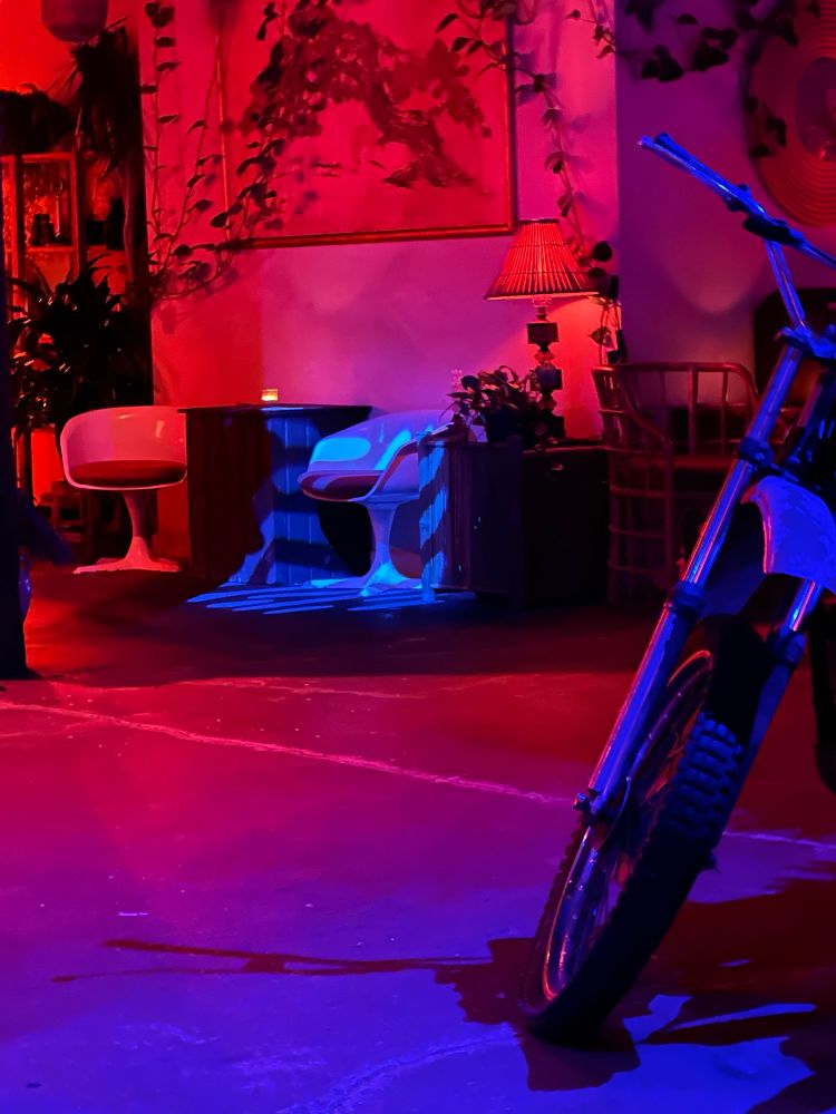 The prompt says not to explain the photo but for the alt text this image has a motorcycle inside of a building with red and blue lighting in the foreground to the right. The background has chairs and other stuff I don’t know I can’t see lol