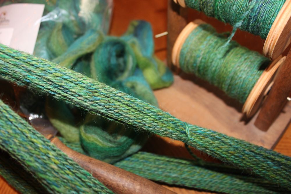 Wool roving, two bobbins of spun yarn, and plied yarn on a Niddy Noddie. It has been dyed in greens and blues.