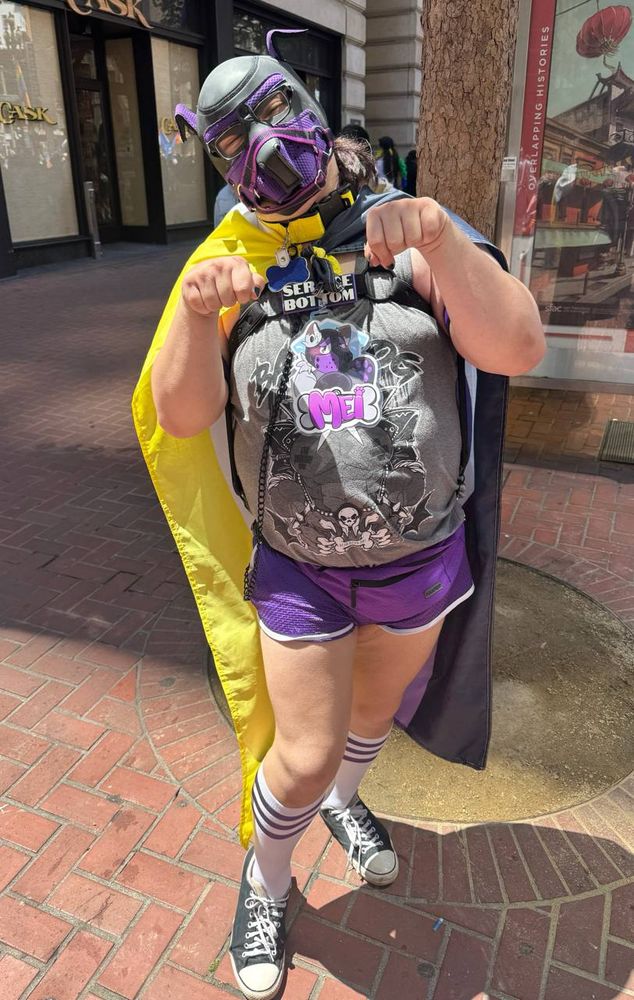 A picture of me, Mei (they/them), posing like a pup in my black and purple pup gear. I’m seen here wearing purple athletic shorts, socks and a fanny pack, as well as a grey graphic tank top with my fursona badge reading “Mei” on top of it. A nonbinary pride flag is also caped around my shoulders.