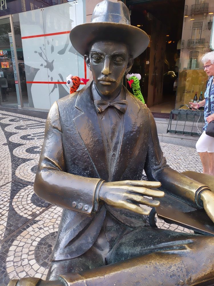 The statue of Fernando Pessoa in Chiado, Lisbon, Portugal. On the shoulders of the legendary Portuguese poet are the two Cobra Meeting mascots, Al T. Cobra and Second-Cousin Cuthbert, both seemingly enthused by the literary giant they have encountered.

A man walks behind the statue with a smartphone attached to headphones. He appears unfazed by the affair.