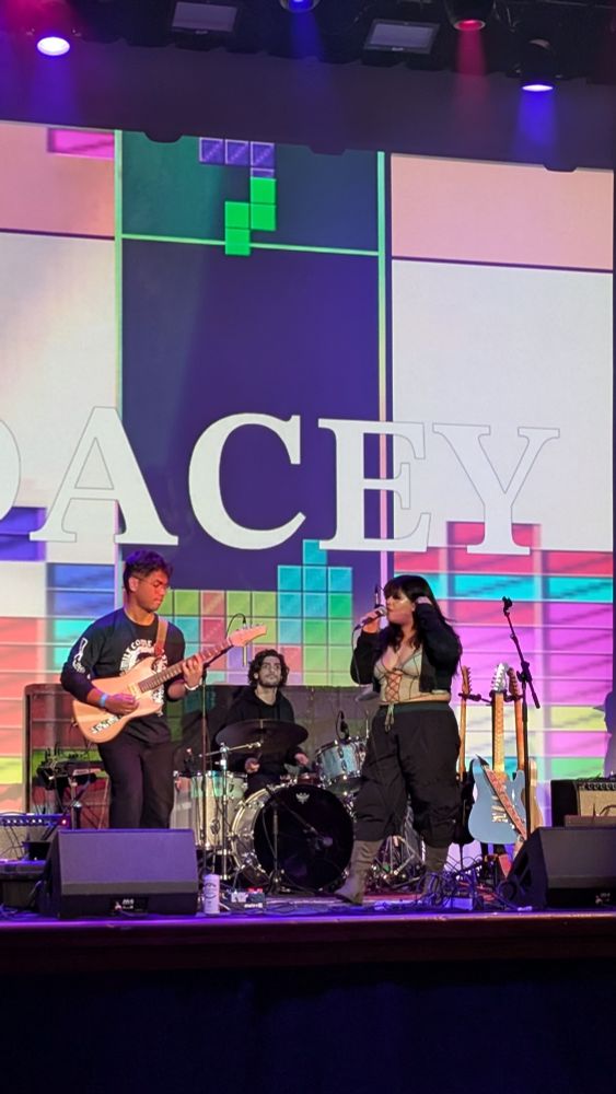 Dacey on stage at the Hollywood Theatre. Behind them is a screen showing their name and a looping game of Tetris. The person playing in the recorded footage is... fine at tetris.