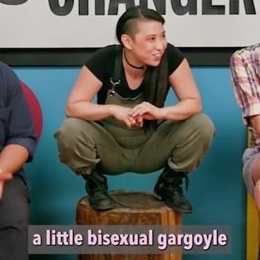 Screenshot from Game Changer with Erika Ishii crouching on a stump like a little bisexual gargoyle (as noted in the closed captions on screen) 